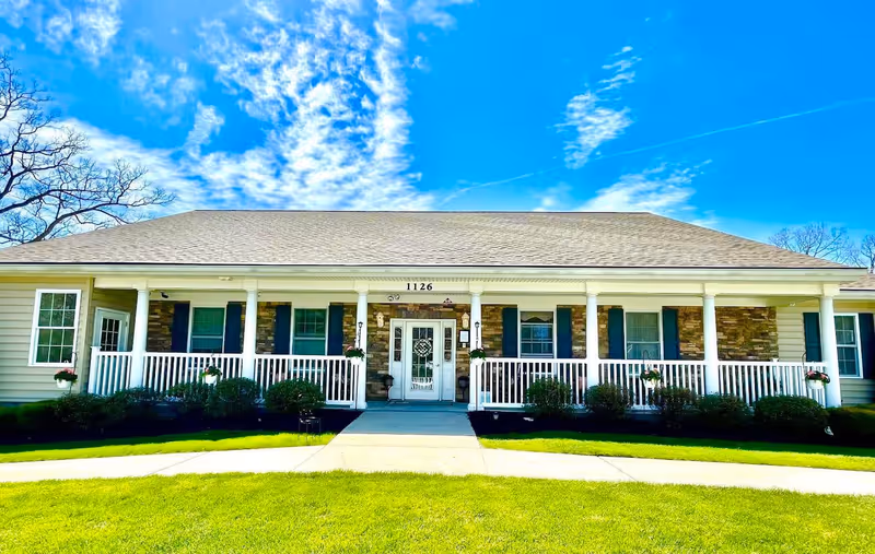 Front exterior view of a single-story building with a light-colored roof, white columns, and a porch with white railings. The building has several windows with dark shutters and hanging flower pots. The address number 1126 is displayed above the entrance door. The sky is bright blue with scattered clouds, and there is green grass and a sidewalk in front of the building.