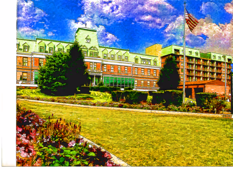 Exterior view of a large senior living facility building with multiple floors, a green roof, and a well-maintained garden with flowers and shrubs in the foreground. An American flag is flying on a flagpole near the building under a partly cloudy blue sky.