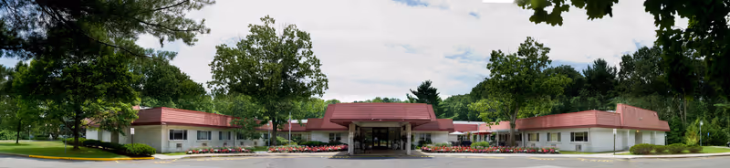 Front exterior view of Autumn Lake Healthcare at Old Bridge, a single-story building with a red roof surrounded by trees and landscaped flower beds under a partly cloudy sky.