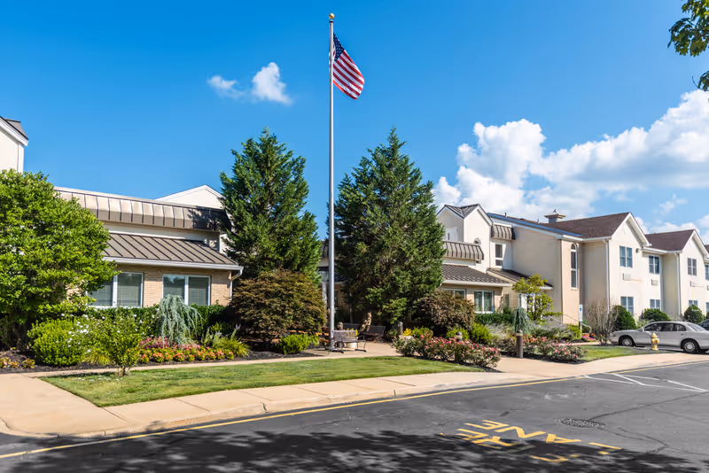 Exterior view of a senior living facility with a well-maintained garden, an American flag on a tall flagpole, and a clear blue sky with some clouds. The building has multiple windows and a beige facade with brown roofing.