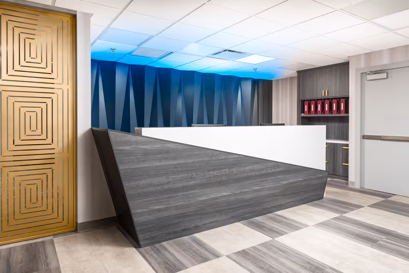 Modern reception desk area with a geometric design, featuring a gray and white desk, a blue-lit textured wall behind it, a gold decorative panel on the left, and gray cabinetry with red binders on the right.