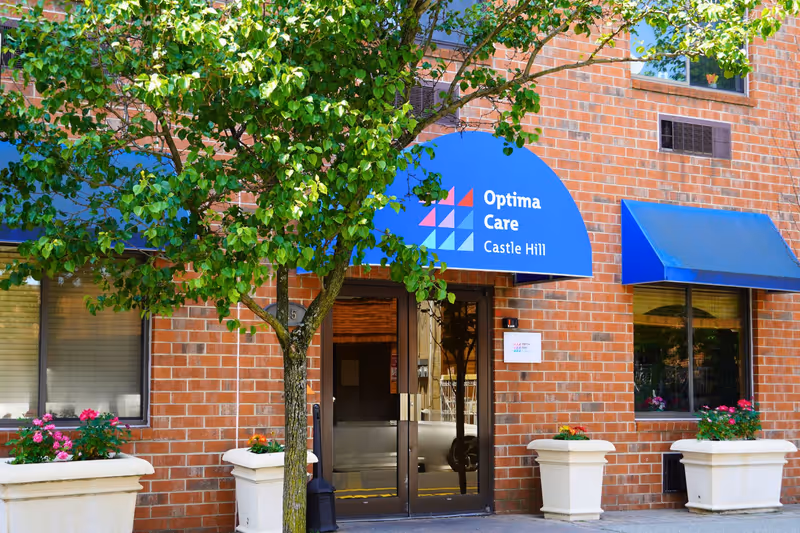 Brick building entrance with a blue awning reading "Optima Care Castle Hill", potted flowers, and a tree in front.