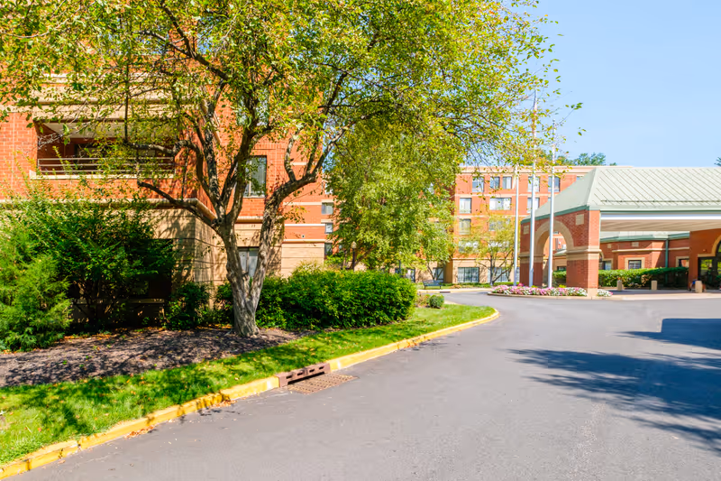 Exterior view of a senior living facility with a paved driveway, green trees, bushes, and a brick building with multiple windows. There is a covered entrance with a green roof and flower beds near the entrance.