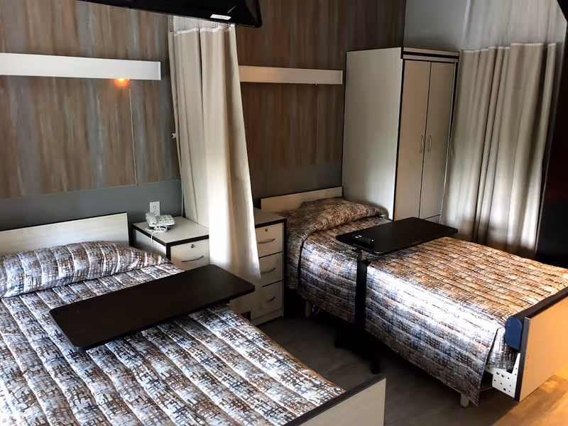 A room with two single beds separated by a curtain. Each bed has a patterned bedspread and an overbed table. There is a small nightstand with a telephone between the beds and a wardrobe in the corner. The room has a neutral color scheme with wood paneling on the wall behind the beds.