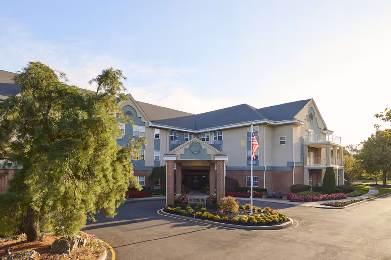 Exterior view of a three-story senior living facility building with a covered entrance supported by brick columns, surrounded by landscaped flower beds and trees, with an American flag on a flagpole in front.