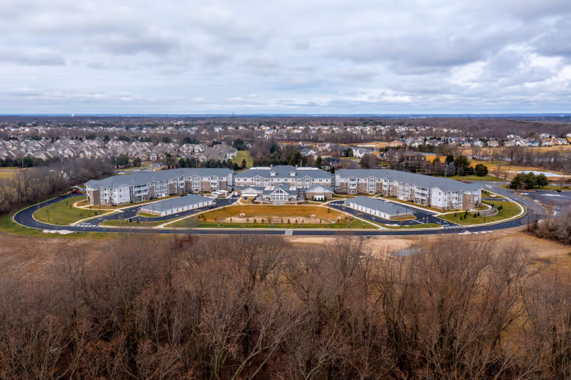 Aerial view of Parkers Bend Retirement Community, a large U-shaped building surrounded by parking lots, roads, and landscaped areas, with a residential neighborhood and trees in the background under a cloudy sky.