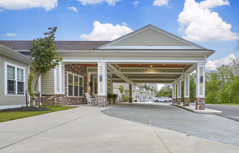 Front exterior view of a senior living facility with a covered drop-off area supported by columns, a paved driveway, landscaped greenery, and several parked cars in the background under a partly cloudy sky.