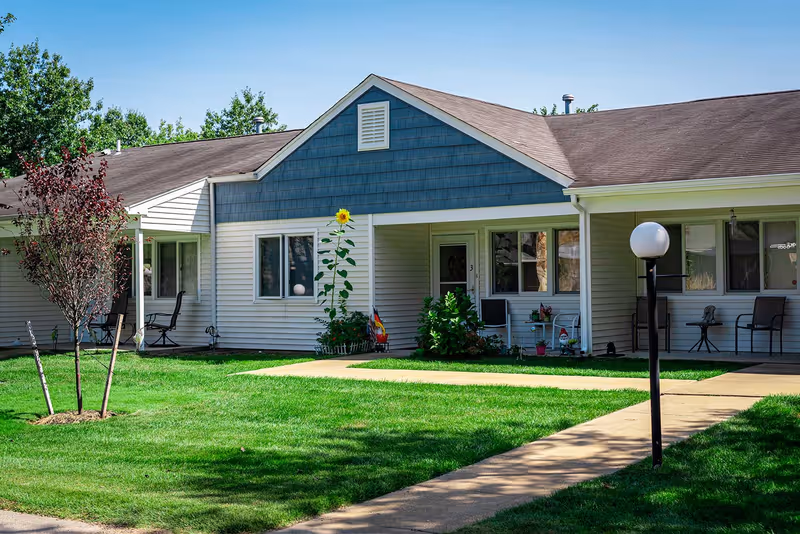 Exterior view of a single-story residential building with white siding and blue gable, featuring a small porch with chairs and a walkway leading to the entrance. There is a well-maintained green lawn with a few small trees and a lamp post in front.