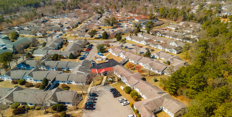 Aerial view of a senior living facility named Silverwoods, showing multiple single-story residential buildings with gray and beige roofs, surrounded by trees and parking lots with several cars parked. The area is well-maintained with clear roads and pathways connecting the buildings.