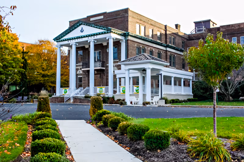 Brick Masonic Village building with a white columned portico, covered driveway and landscaped walkway in front.