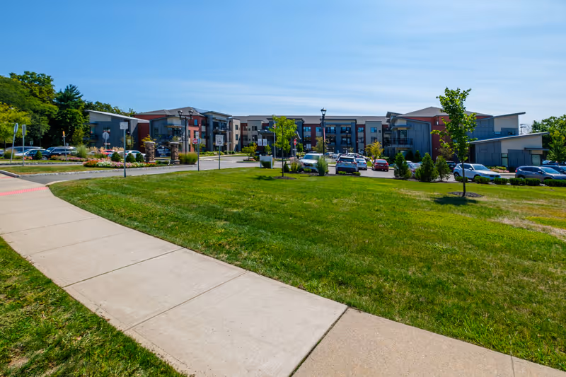 Wide exterior view of Brightview Wayne senior living facility on a sunny day, showing a modern multi-story building with balconies, a parking lot with cars, green lawn, trees, and a sidewalk in the foreground.
