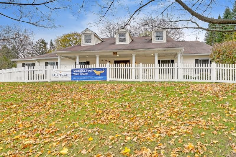 Exterior view of Fox Trail Memory Care Living facility, a single-story building with a brown roof and white siding, surrounded by a white picket fence. The lawn in front is covered with fallen autumn leaves, and there are trees in the background. A banner on the fence invites visitors to schedule a visit.