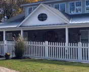 Front exterior view of a residential-style building with white siding, stone accents, a metal roof, and a white picket fence in front. There is a small oval window above the entrance and some plants near the fence.