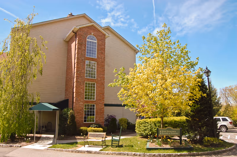 Exterior view of a multi-story assisted living facility building with beige siding and a central brick section featuring tall windows. The scene includes a green canopy over an entrance, a bench, a rocking chair, trees with green and yellow leaves, a lamp post, and a sign indicating the main entrance and handicap parking. A white SUV is parked in the background under a clear blue sky.