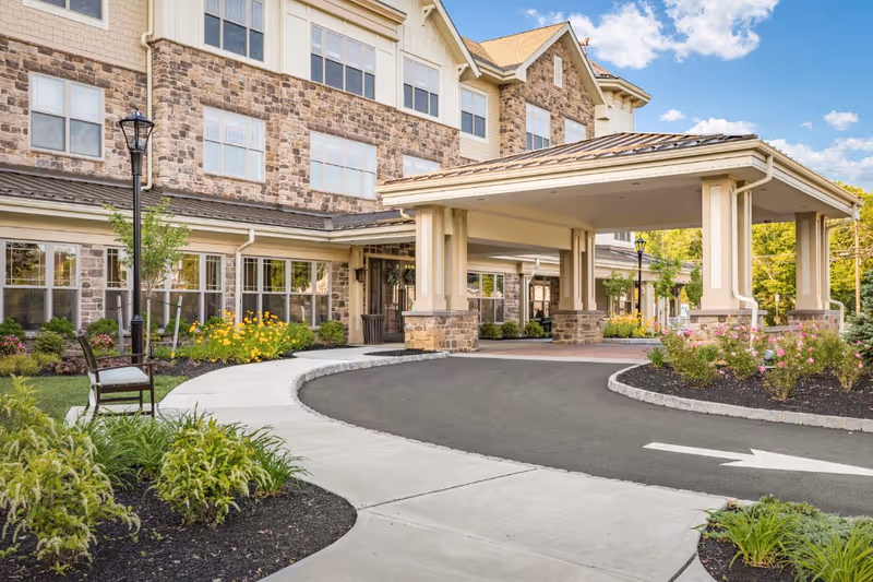 Covered porte-cochère entrance and landscaped circular driveway of a senior living building with a stone facade.