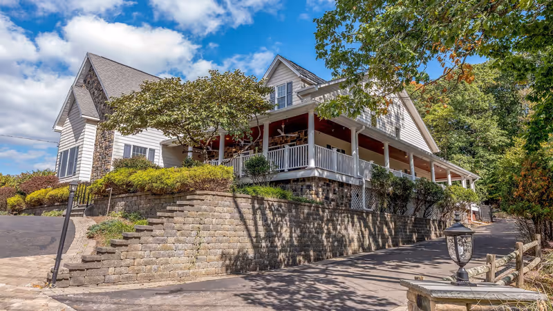 Exterior view of a senior living facility building with white siding, stone accents, and a large covered porch with white railings. The building is surrounded by greenery, trees, and a stone retaining wall along a paved driveway under a partly cloudy blue sky.