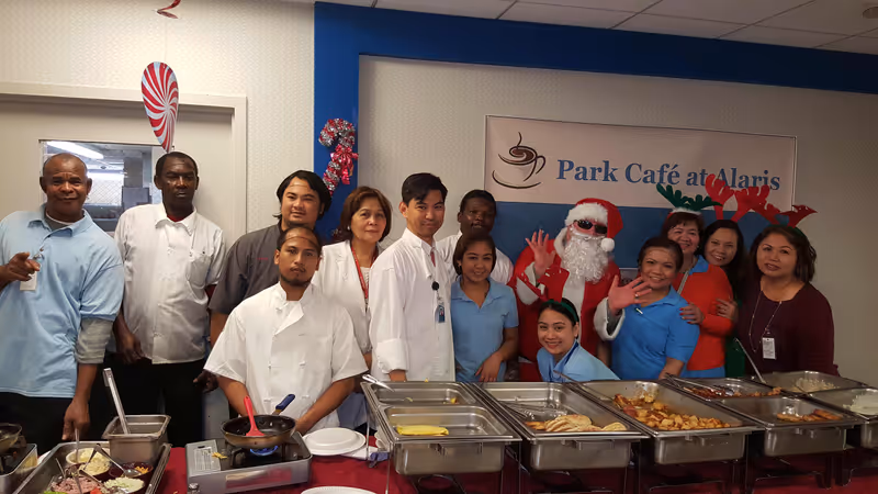 A group of staff members and a person dressed as Santa Claus posing behind a buffet table with various food trays in a room decorated for the holidays at Park Café at Alaris. Some people are wearing festive accessories like reindeer antlers and Santa hats.