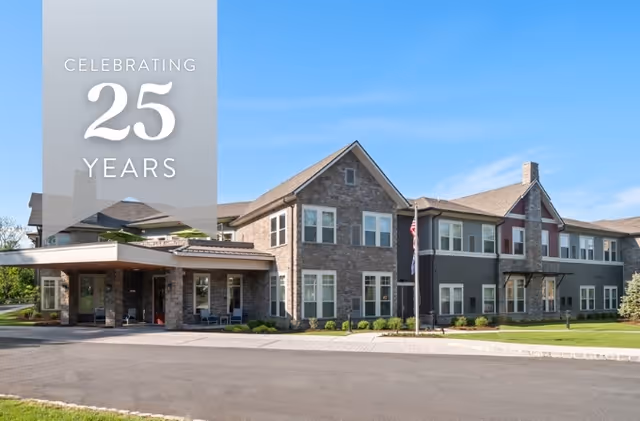 Exterior view of The Bristal Assisted Living at Waldwick building on a clear day with a blue sky. The building has a combination of stone and siding facade with multiple windows and a covered entrance. There is a flagpole with an American flag in front of the building and well-maintained landscaping along the sidewalk.