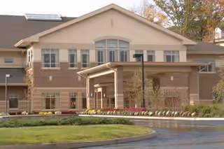 Exterior view of a two-story senior living facility building with a covered entrance, multiple windows, and landscaped grounds including grass and flower beds.