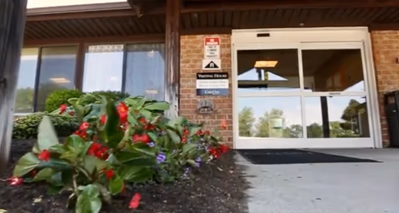 Entrance to a building with glass automatic sliding doors, a brick exterior wall, and a flower bed with red and purple flowers in the foreground.