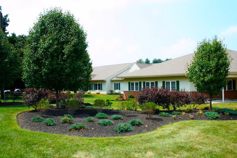 A single-story building with beige siding and green shutters surrounded by a well-maintained garden with various shrubs, small trees, and a neatly trimmed lawn under a partly cloudy sky.