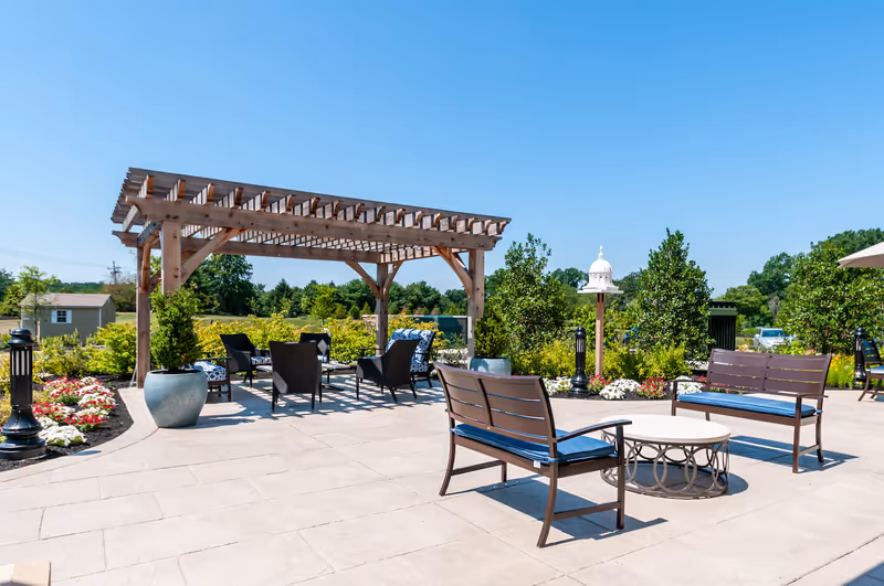 Outdoor patio area with a wooden pergola, several cushioned chairs, benches with blue cushions, a round table, potted plants, flower beds, and a birdhouse on a post under a clear blue sky.