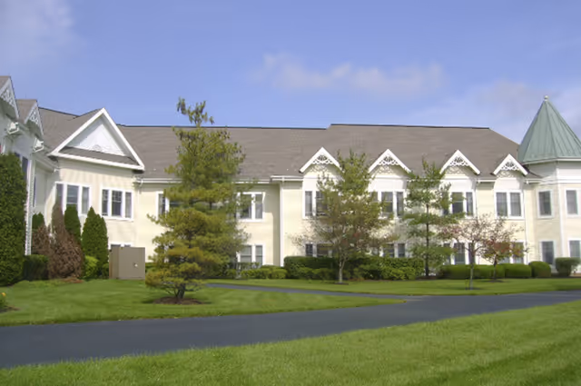 Exterior view of a two-story senior living facility building with cream-colored siding, multiple windows, decorative gables, and a green conical roof tower. The building is surrounded by a well-maintained lawn, small trees, and shrubs under a partly cloudy sky.