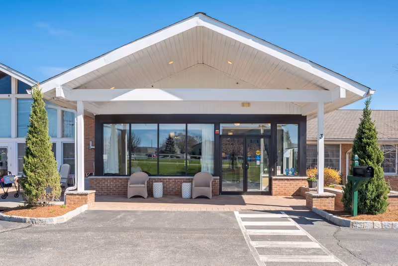 Front entrance of an assisted living facility with a covered porte-cochère, glass doors, two chairs, and landscaping.