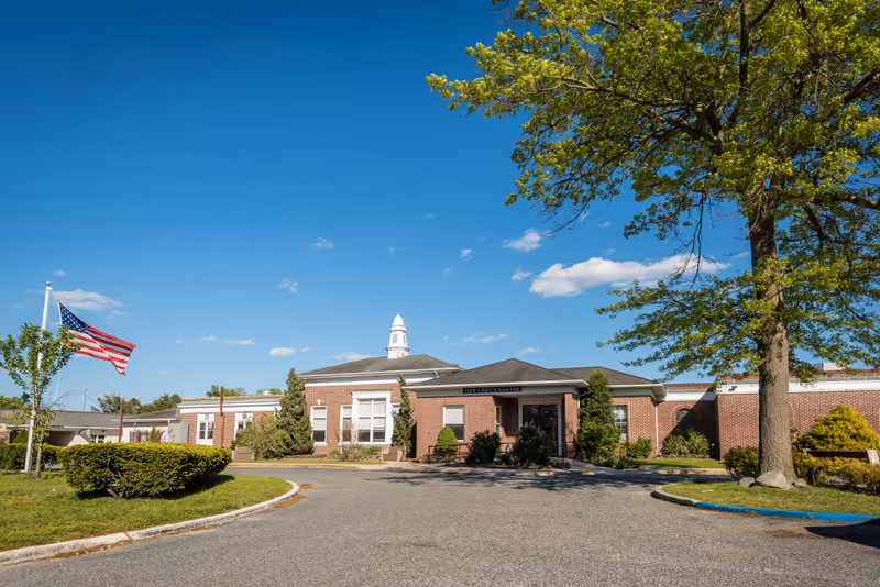 Front exterior view of Our Lady's Center For Rehabilitation & Healthcare, a single-story brick building with a white cupola on the roof, surrounded by greenery and trees under a clear blue sky. An American flag is flying on a flagpole to the left.
