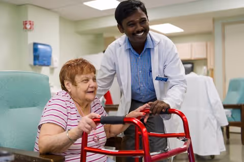 An elderly woman sitting in a chair using a red walker, smiling, with a male healthcare professional in a white coat standing beside her, also smiling, in a well-lit room with medical and office furniture in the background.