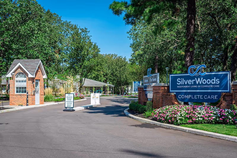 Entrance to SilverWoods senior living facility with a brick guardhouse on the left, a paved driveway, and a large blue sign on the right that reads 'SilverWoods Independent Living by Complete Care' surrounded by flowers and trees under a clear blue sky.