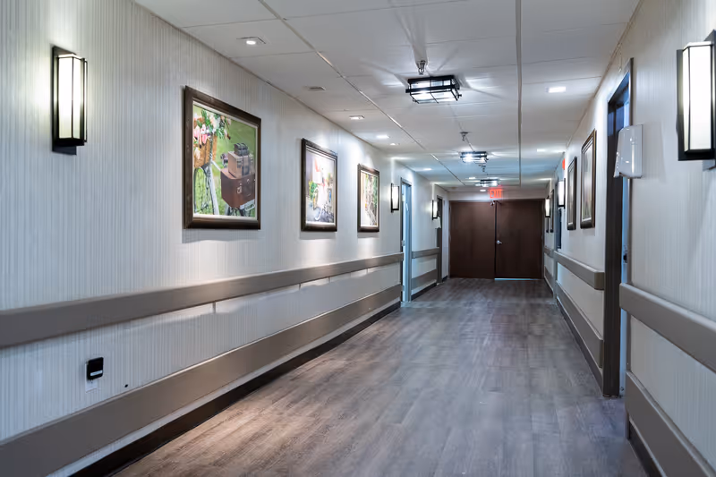 A long, well-lit interior hallway with framed artwork on the walls, handrails, and double doors at the far end.