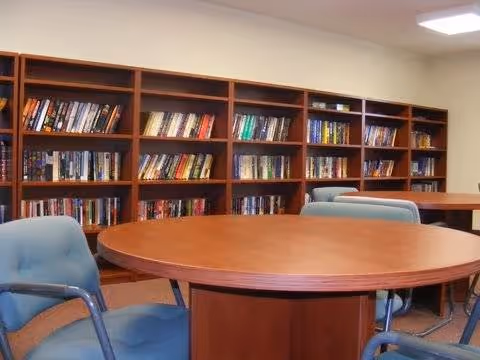 Interior view of a room with wooden bookshelves filled with books along the wall, a round wooden table in the center, and blue cushioned chairs around the table.