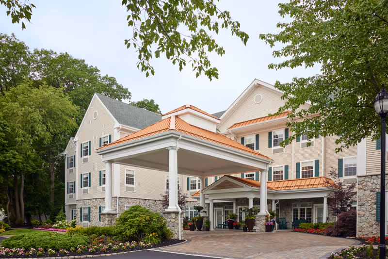 Exterior view of a senior living facility building with beige siding, green shutters, and a stone foundation. The entrance features a covered porte-cochère with white columns and a copper-colored roof. There are landscaped flower beds and trees surrounding the driveway leading to the entrance.