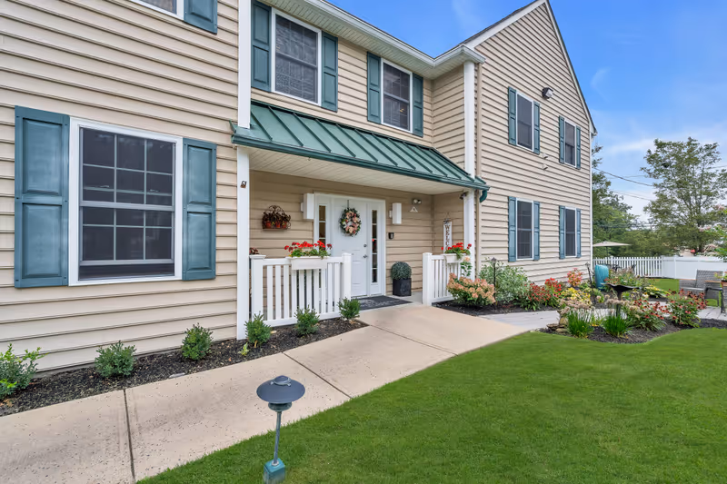 Exterior view of a beige two-story building with green shutters and a green metal awning above the white double front doors. There is a small porch with white railings, flower pots with red flowers, and a welcome sign. The building is surrounded by a well-maintained lawn, flower beds, and a paved walkway leading to the entrance.