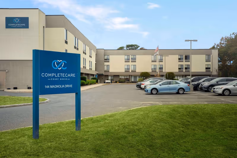 Exterior view of Complete Care at Court House building with a parking lot containing several cars. A blue sign in the foreground displays the facility name and address: 144 Magnolia Drive. The building is three stories tall with multiple windows and an American flag near the entrance.