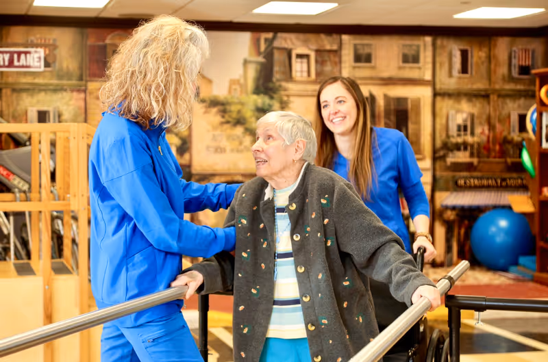 An elderly woman using parallel bars for walking assistance is supported by two female caregivers wearing blue uniforms in a bright, indoor therapy or rehabilitation area with a mural of buildings in the background.